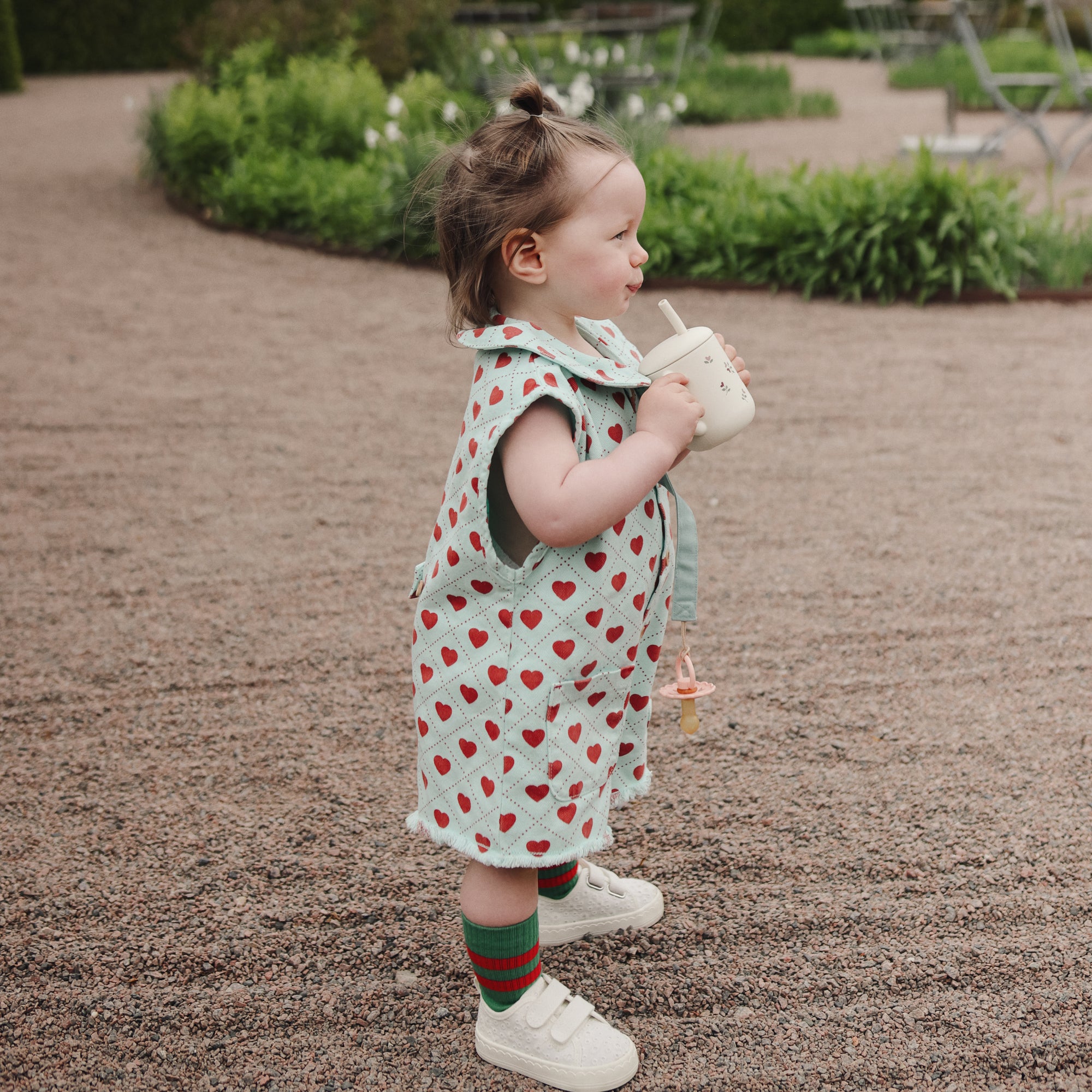 Toddler wears mushie Linen Pacifier Clip 2-Pack while drinking outdoors in a heart-print outfit.
