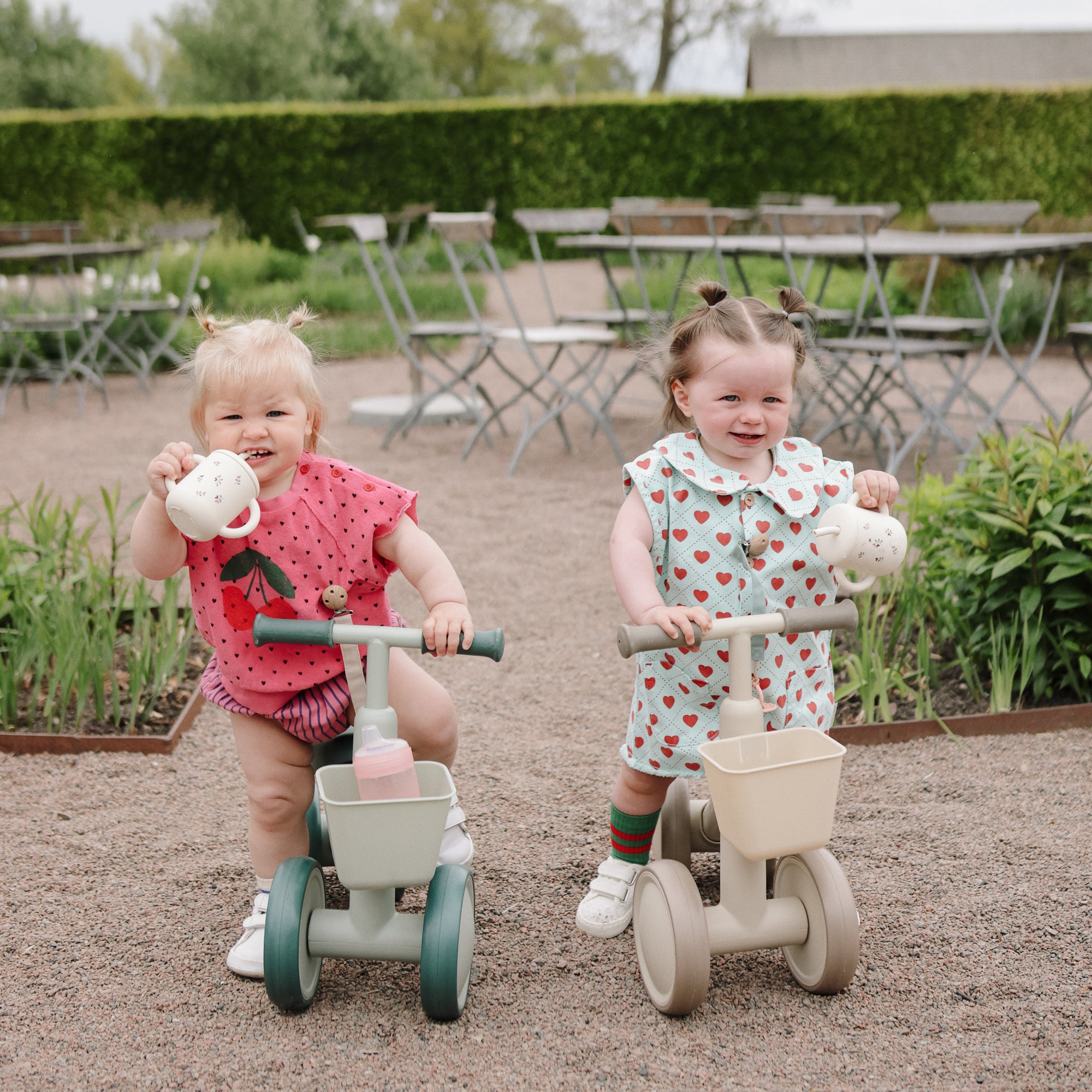 Two toddlers ride mushie Mini Toddler Bikes outdoors, smiling and holding toy teapots.