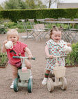 Two toddlers ride mushie Mini Toddler Bikes outdoors, smiling and holding toy teapots.