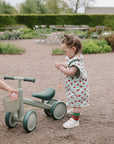 Two toddlers ride a green mushie Mini Toddler Bike on a gravel path in the garden.