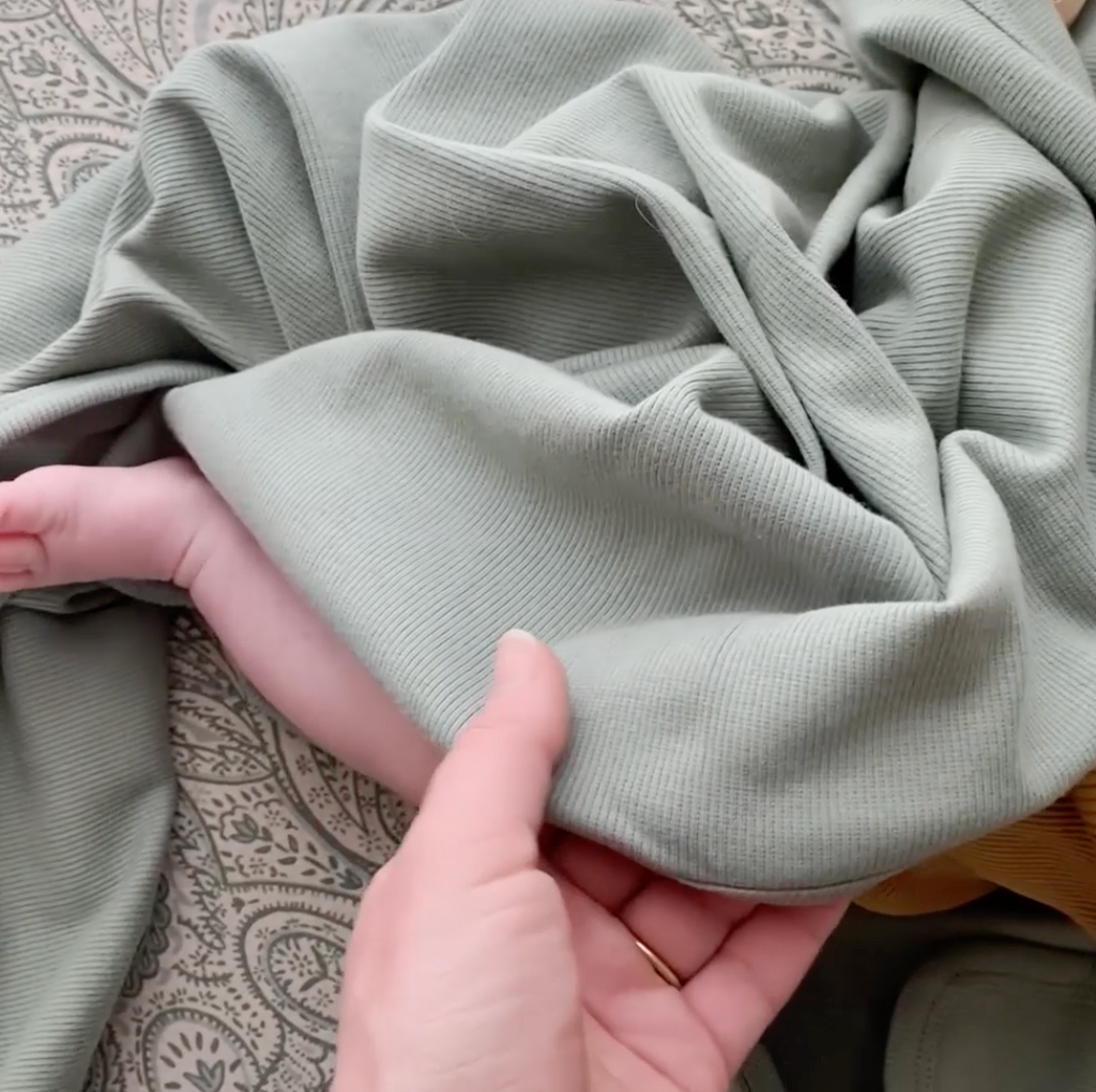 A behind the scenes sneak peek of the mushie team designing the blanket in a white room, comparing swatches of cloth, then transitioning to seeing the full ribbed baby blanket with a little baby. 