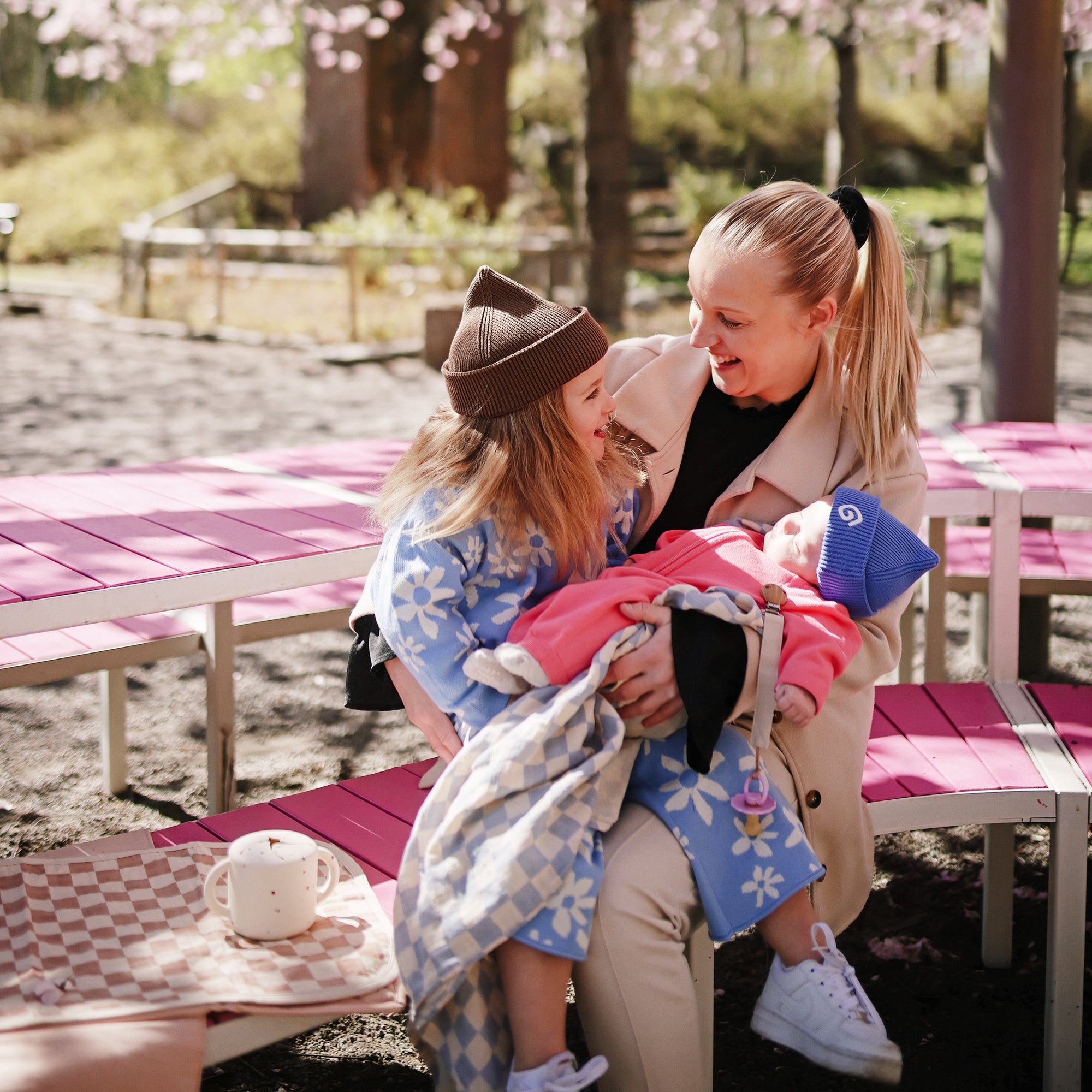 A smiling woman holds a baby with a FRIGG Daisy Natural Rubber Pacifier by pink benches.