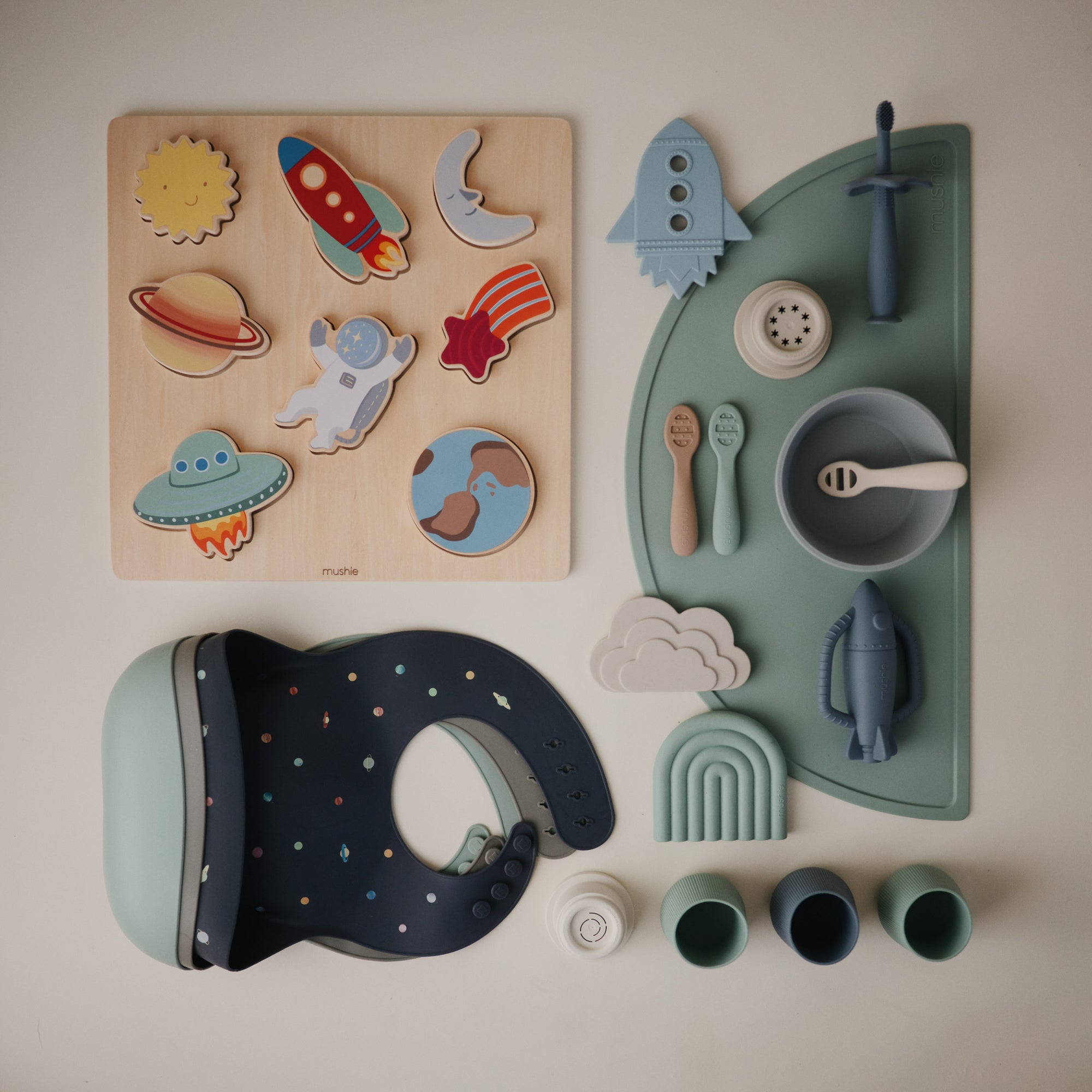 A set of baby items with a space theme, including a space-themed wooden puzzle, silicone bib, bowls, utensils, cups, and a plate shaped like a rocket, all in muted blue, green, and neutral colors on a white surface.