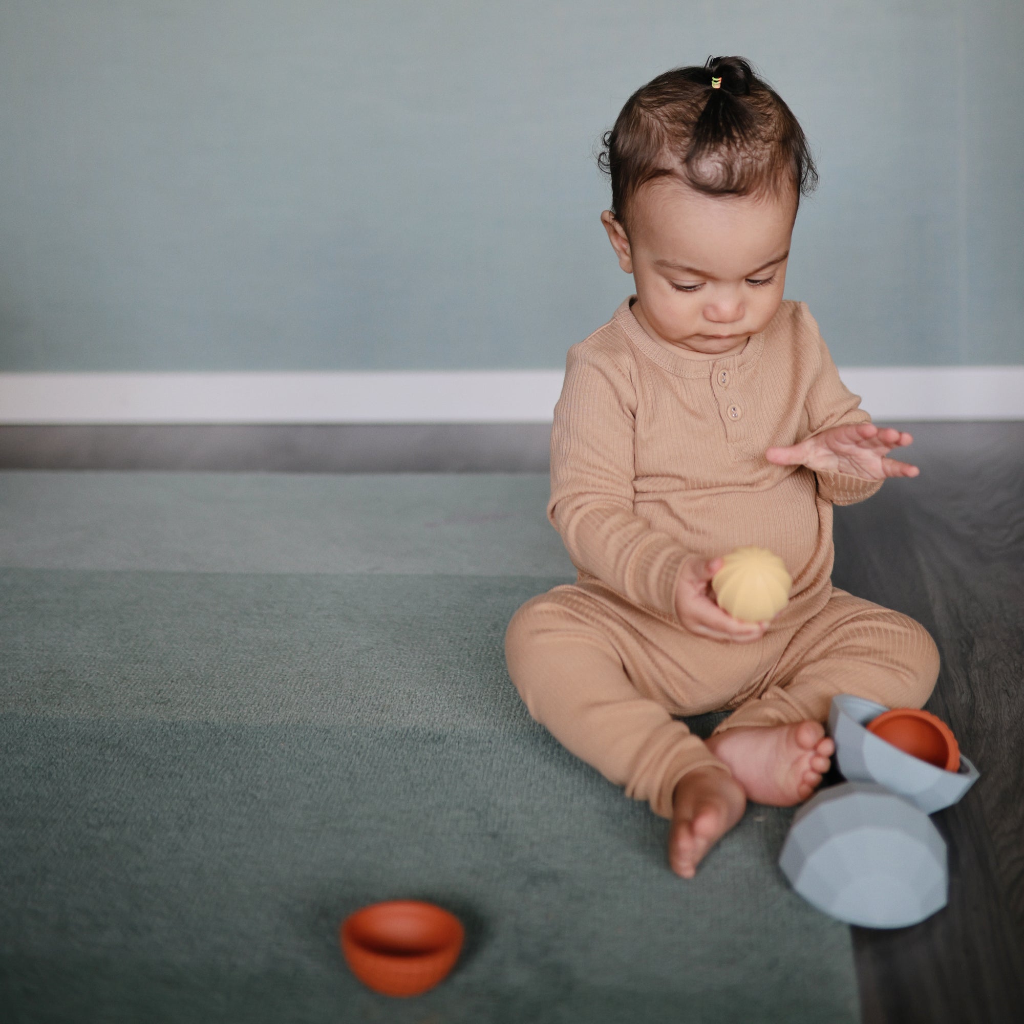 Baby in beige on rug plays with yellow ball and mushie Nesting Spheres Sensory Toy.
