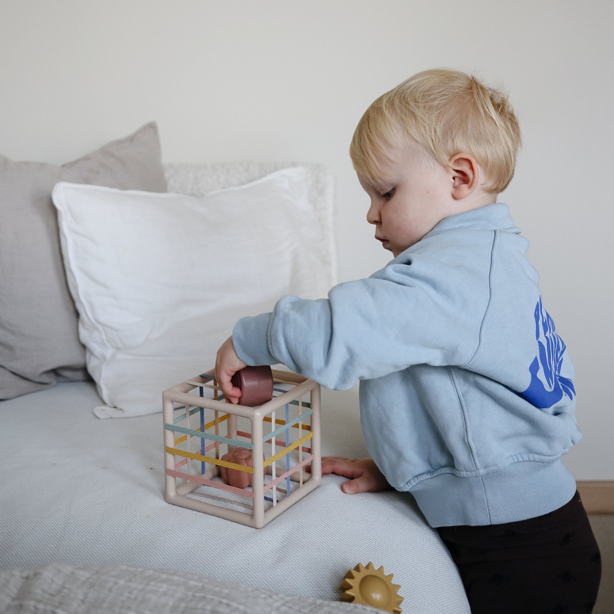 Toddler plays with mushie Elastic Shape Sorter, building fine motor skills by the bed.