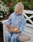 A child sits on a bench holding an open mushie Lunchbox with a spoon.