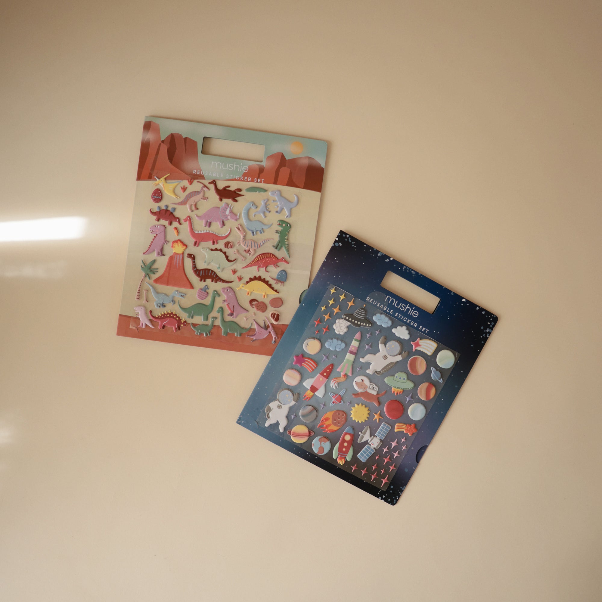 Two packs of mushie sticker sets on a light beige surface: one with dinosaur puffy stickers against a desert background, and the other is a reusable space-themed sticker set featuring astronauts, planets, and rockets on a starry backdrop.