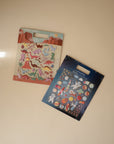 Two packs of mushie sticker sets on a light beige surface: one with dinosaur puffy stickers against a desert background, and the other is a reusable space-themed sticker set featuring astronauts, planets, and rockets on a starry backdrop.