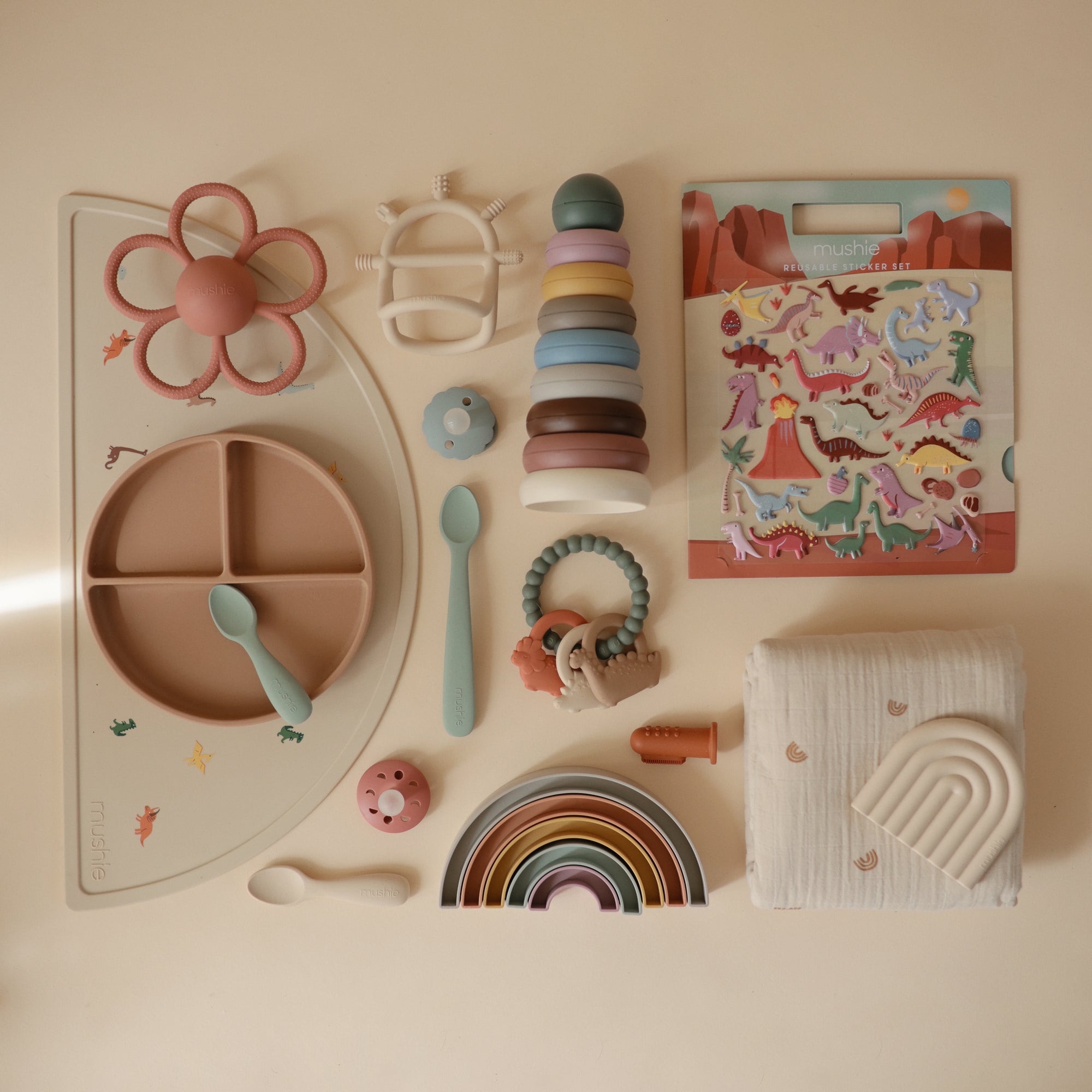 A flat lay of baby items in soft pastels, featuring a divided silicone plate, silicone utensils, stacking toys, teether, pacifier, rainbow toy, muslin cloth, soft textured toy, and the mushie Reusable Sticker Set.