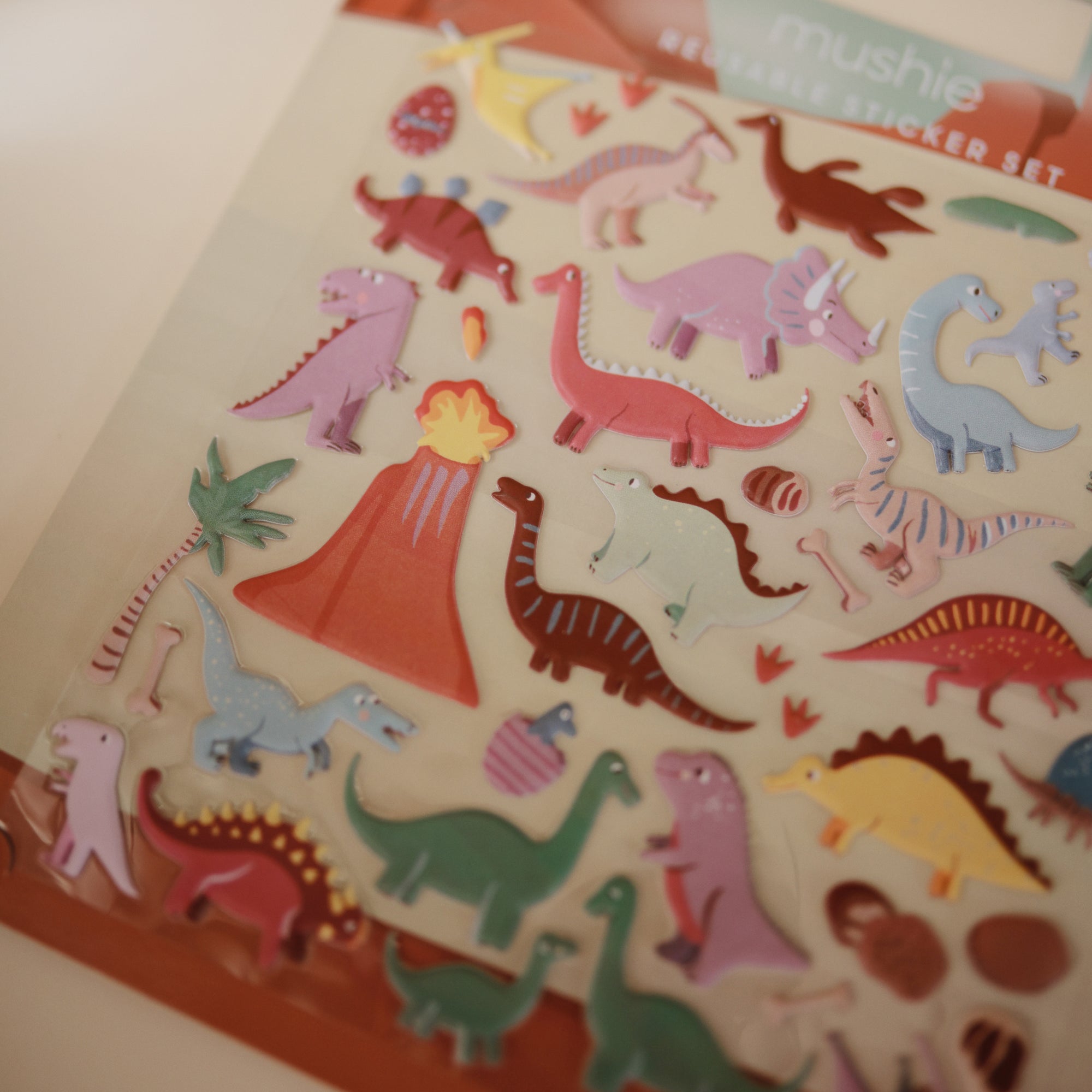 The mushie Reusable Sticker Set features colorful cartoon dinosaurs, a volcano, palm trees, and dino eggs on a beige background—perfect for fans of reusable sticker sets or sticker books.