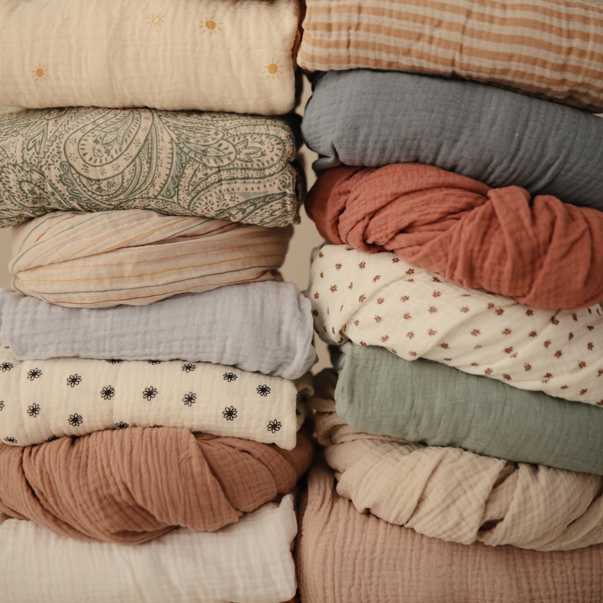 Stack of mushie Mini Muslin Crib Sheets in patterned and solid earthy, neutral hues.