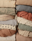 Stack of mushie Mini Muslin Crib Sheets in patterned and solid earthy, neutral hues.