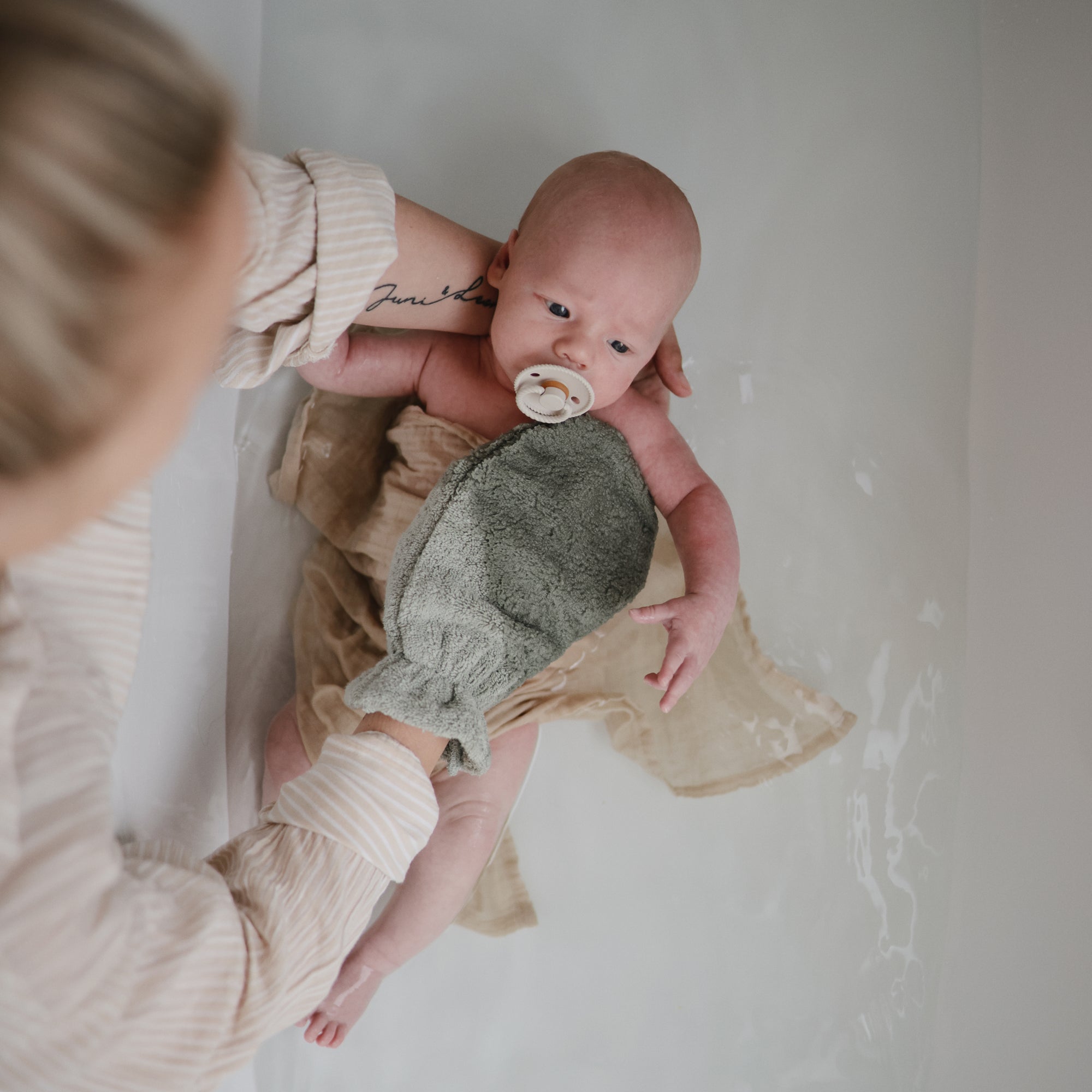 A caregiver bathes a baby in a tub, holding them with one arm while gently washing with a green cloth. The baby calms with the Mushie FRIGG Rope Natural Rubber Pacifier 2-Pack—one of many trusted baby essentials.