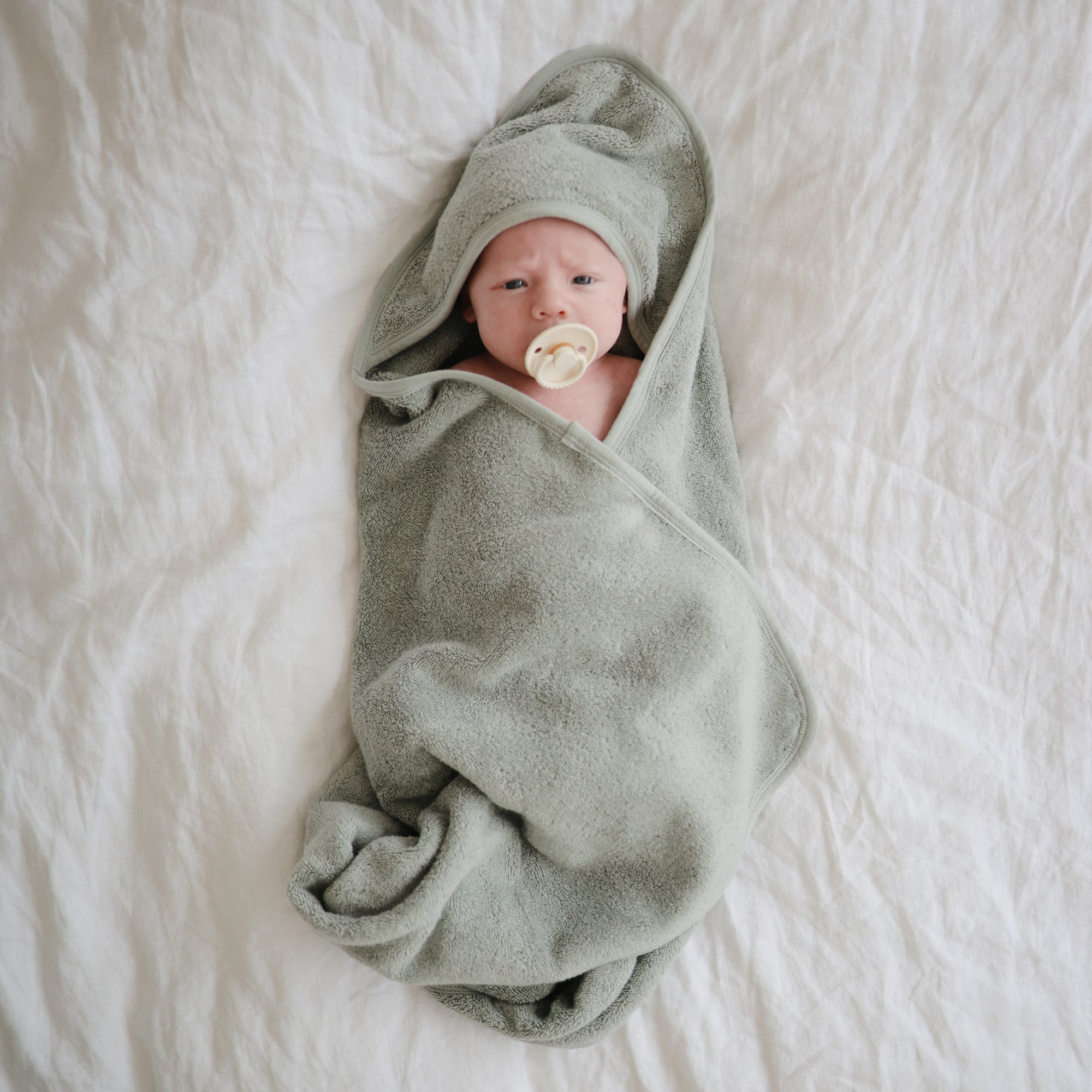 A baby wrapped in a soft, light green hooded towel lies on a white bed, soothing with a Mushie FRIGG Rope Natural Rubber Pacifier—a must-have among baby essentials.