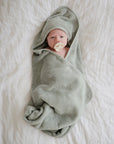 A baby wrapped in a soft, light green hooded towel lies on a white bed, soothing with a Mushie FRIGG Rope Natural Rubber Pacifier—a must-have among baby essentials.
