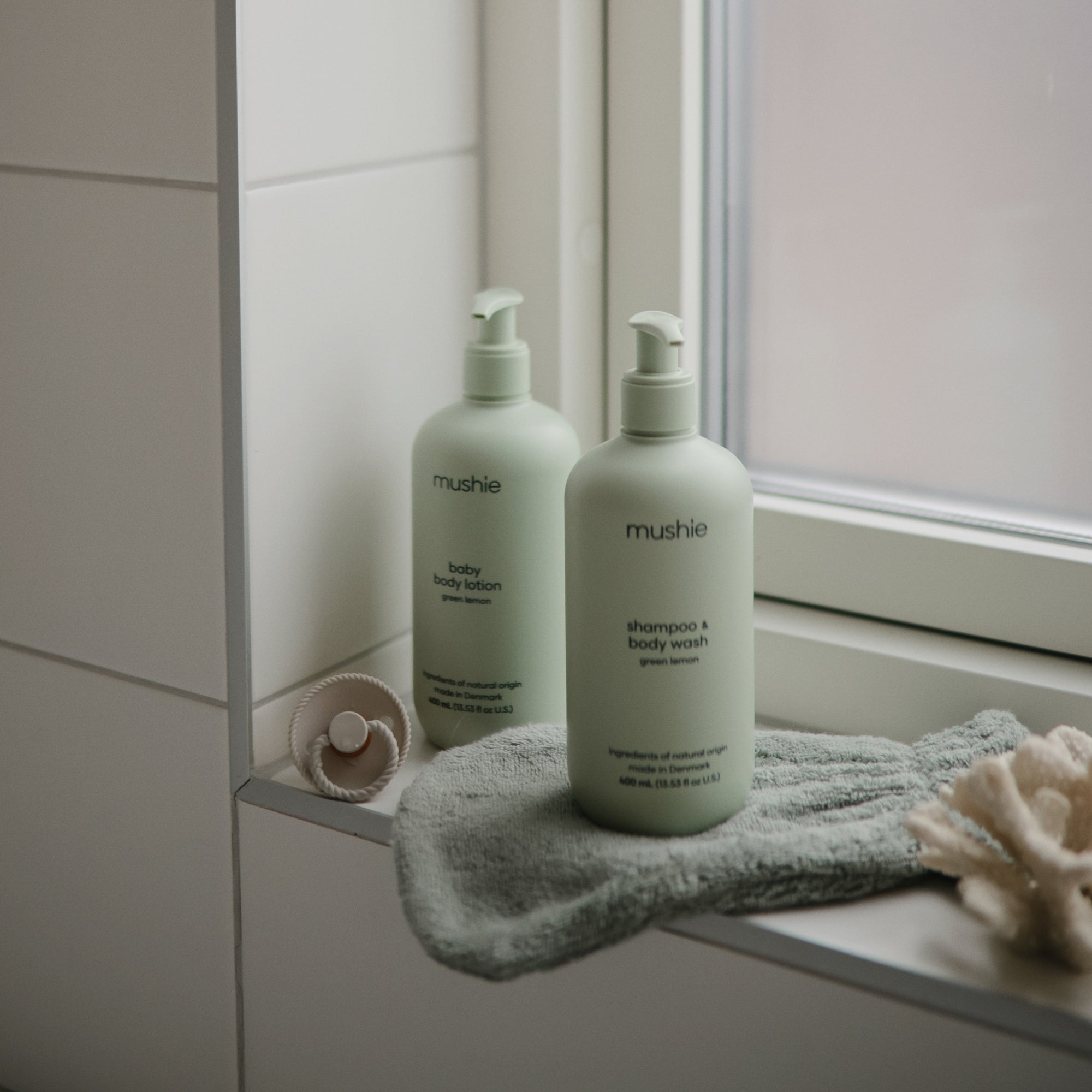Two light green bottles of Mushie baby body lotion and shampoo &amp; body wash sit on a windowsill with baby essentials. They pair perfectly with Mushie's FRIGG Rope Natural Rubber Pacifier 2-Pack.