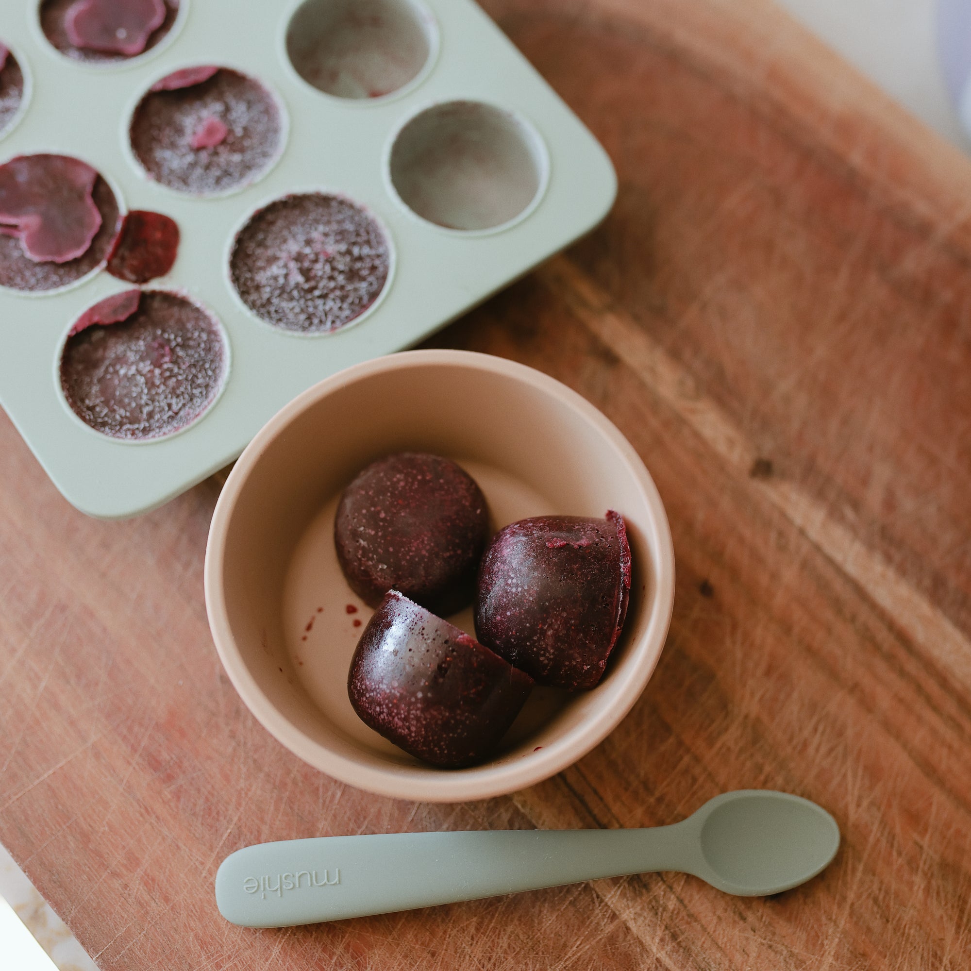 Three berry cubes in a Mushie Baby Food Freezer Tray with baby spoon on wood.