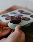 Hands removing a frozen berry mix from a Mushie Baby Food Freezer Tray on a wooden surface.