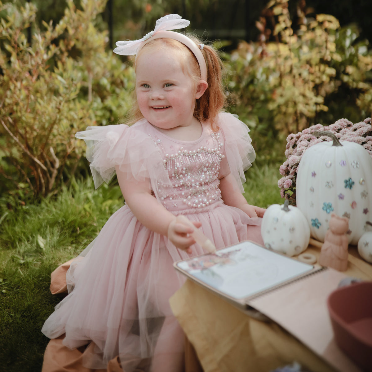 Child in pink dress with pumpkins &amp; mushie Reusable Water Color Pad sits outdoors, smiling.