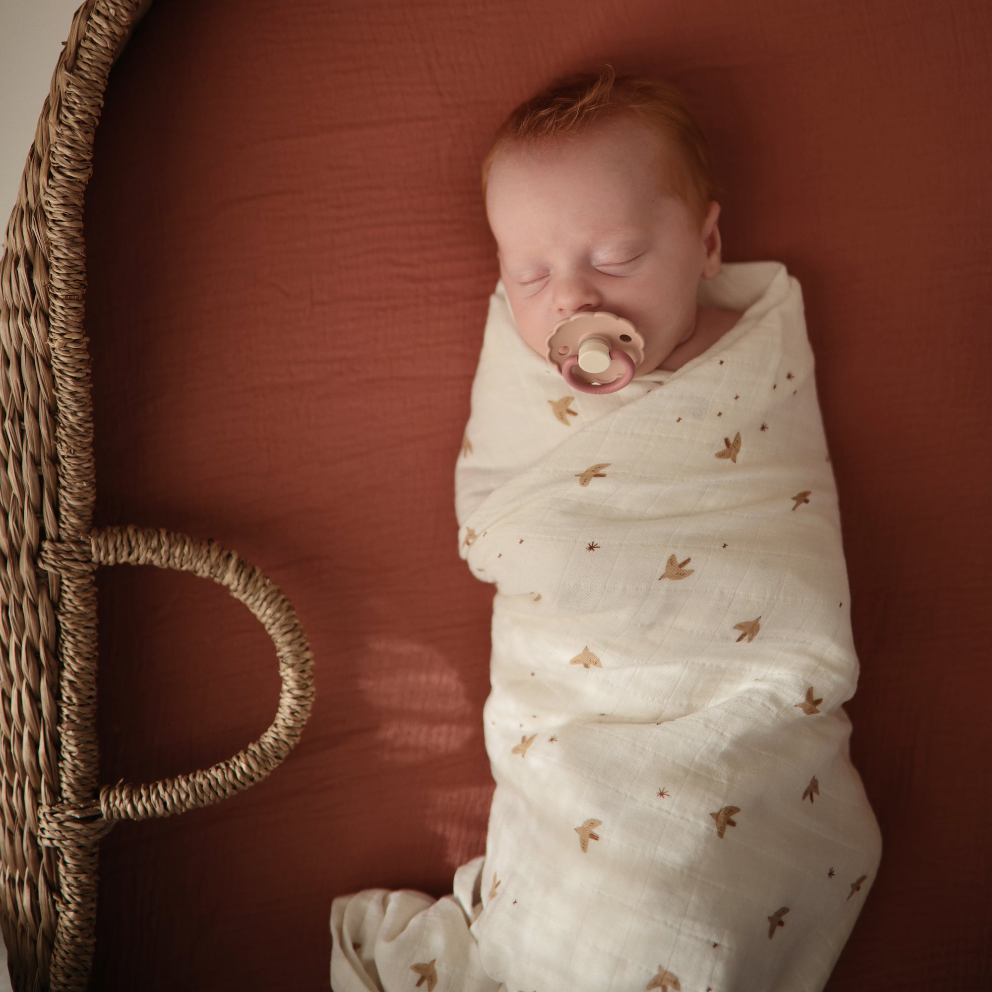 Newborn wrapped in mushie Organic Cotton Muslin Swaddle Blanket, lying in a basket bassinet.
