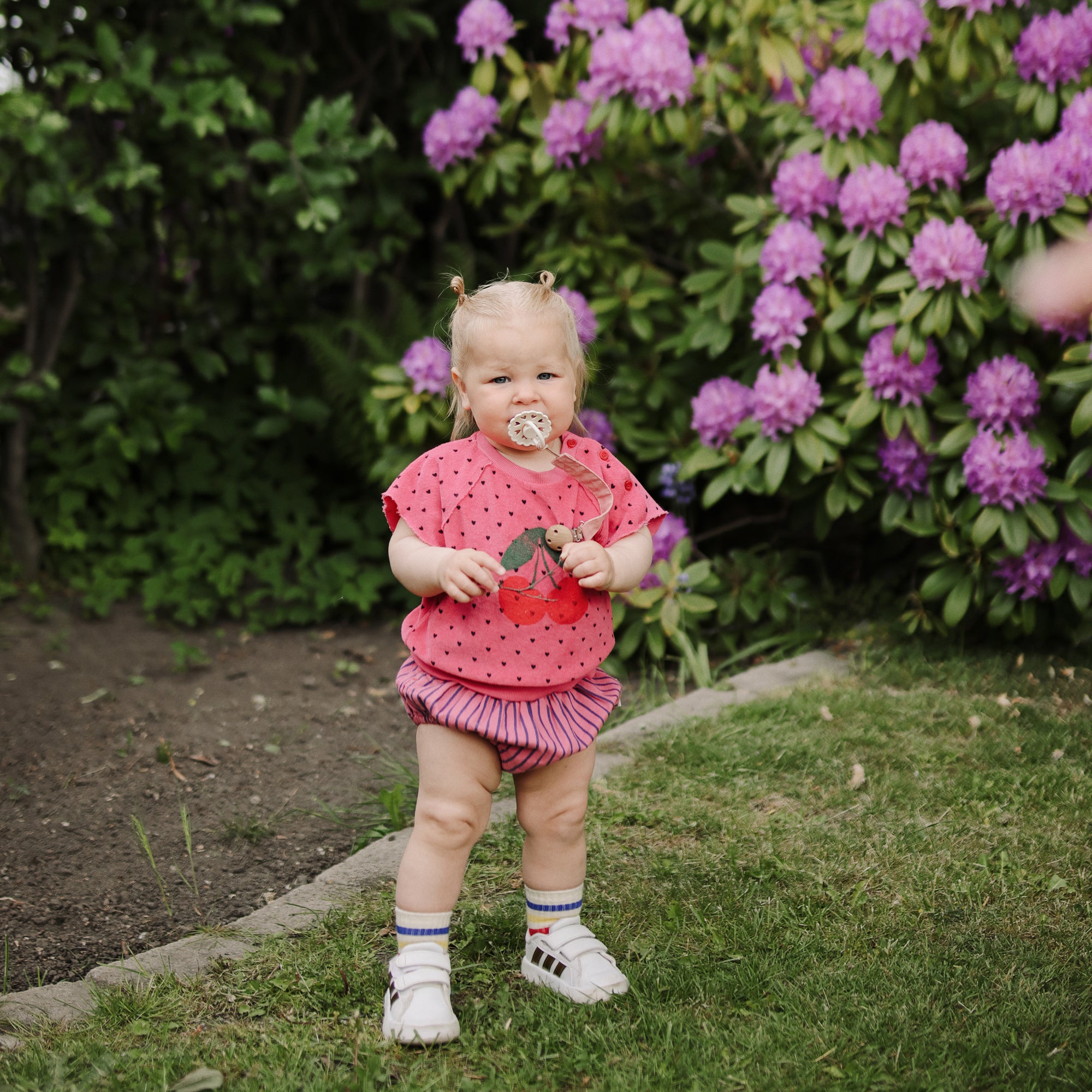 Baby with FRIGG Andersen Fairytale pacifier stands on grass by purple flowers, in pink outfit.