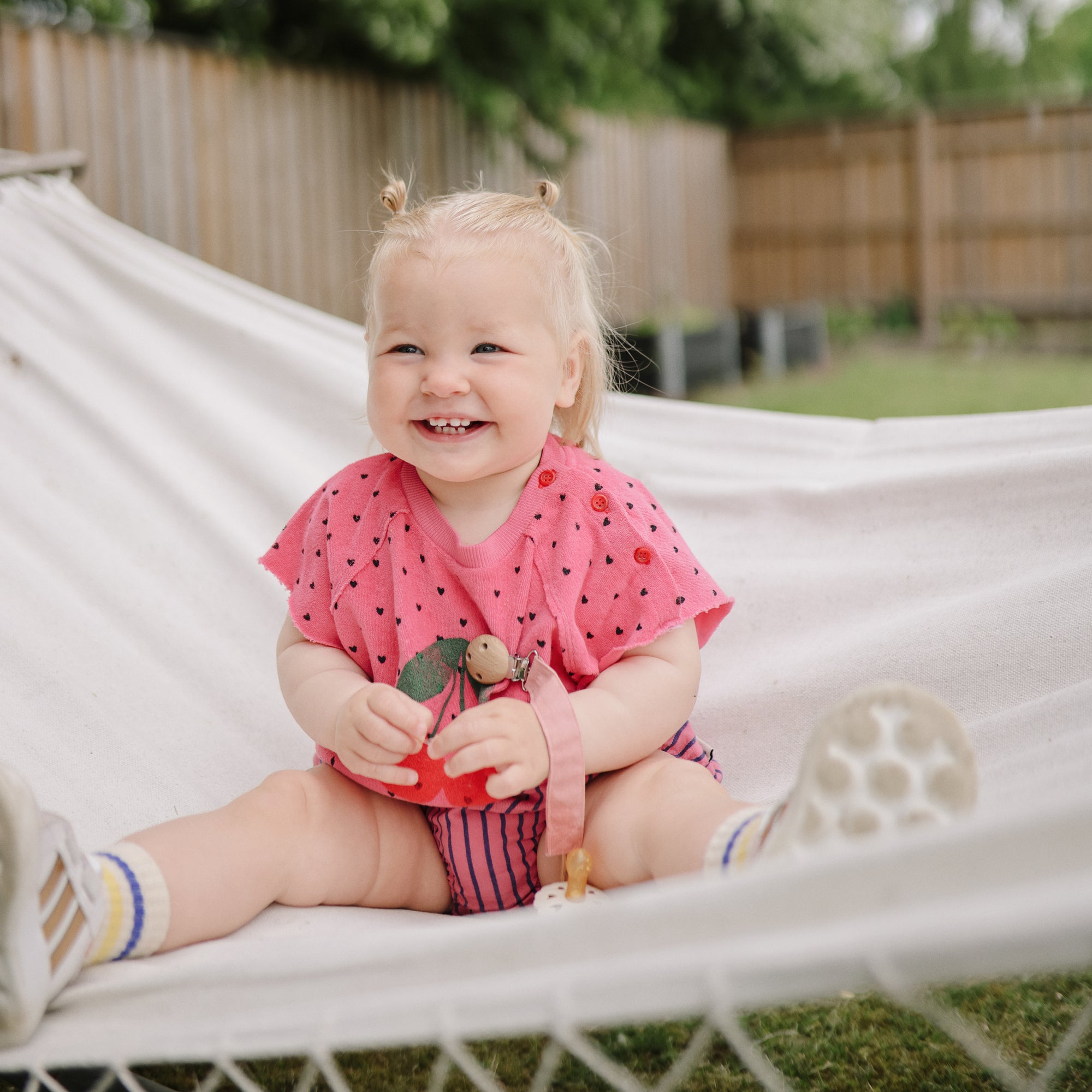 Smiling toddler wears mushie Linen Pacifier Clip 2-Pack on a white hammock outdoors.