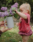 Toddler outdoors with Mushie Bath Rinse Cup, reaching for lilacs in a white vase.