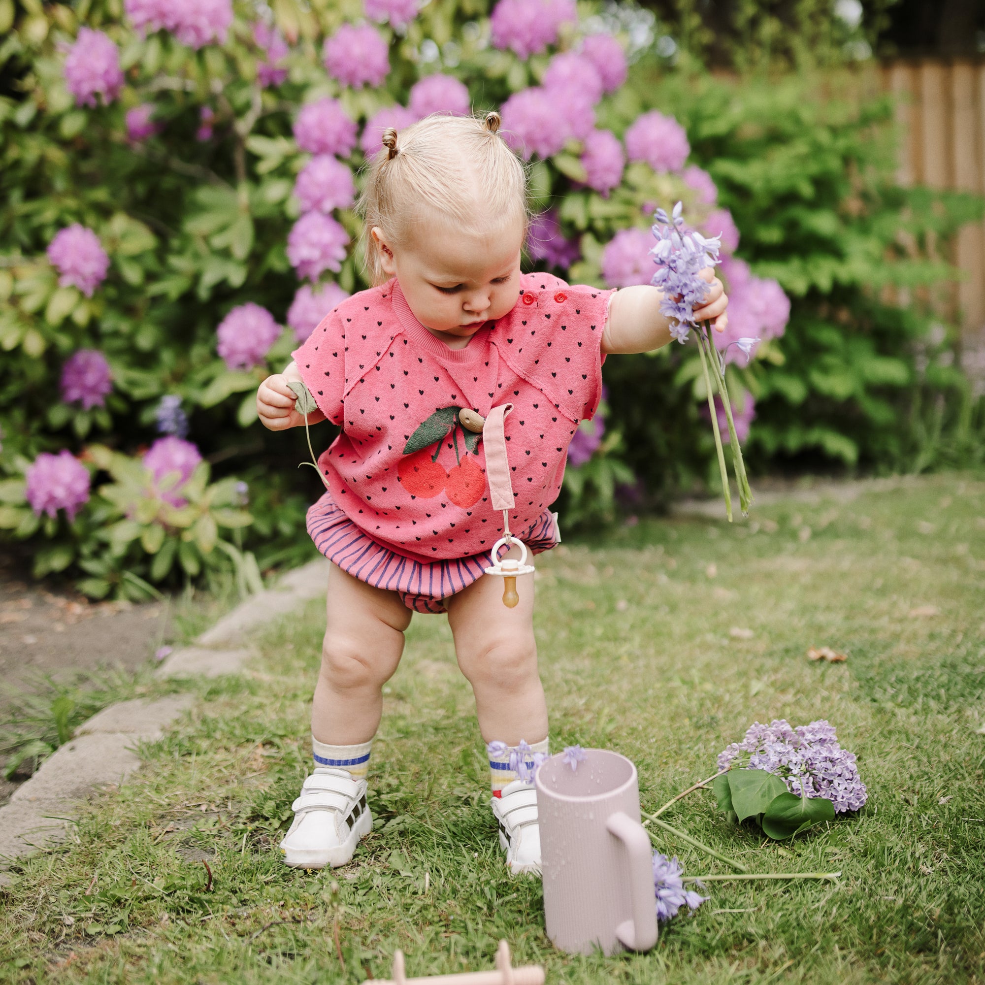 A toddler plays with a Mushie Bath Rinse Cup among lilac and purple flowers.