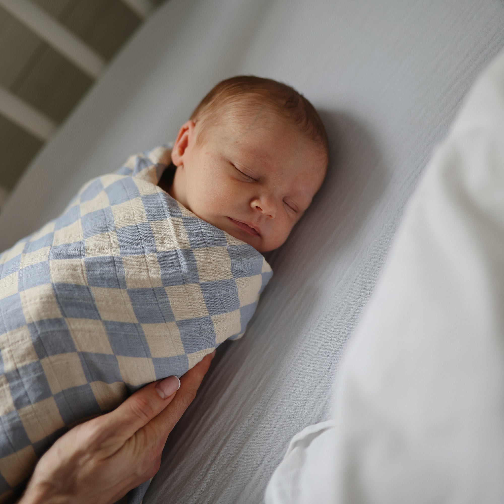 A newborn sleeps soundly, swaddled in a blue and white checkered blanket. An adult hand gently rests on the baby, who lies on mushie Extra Soft Muslin Crib Sheet—breathable, cozy bedding for cribs.