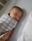 A newborn sleeps soundly, swaddled in a blue and white checkered blanket. An adult hand gently rests on the baby, who lies on mushie Extra Soft Muslin Crib Sheet—breathable, cozy bedding for cribs.