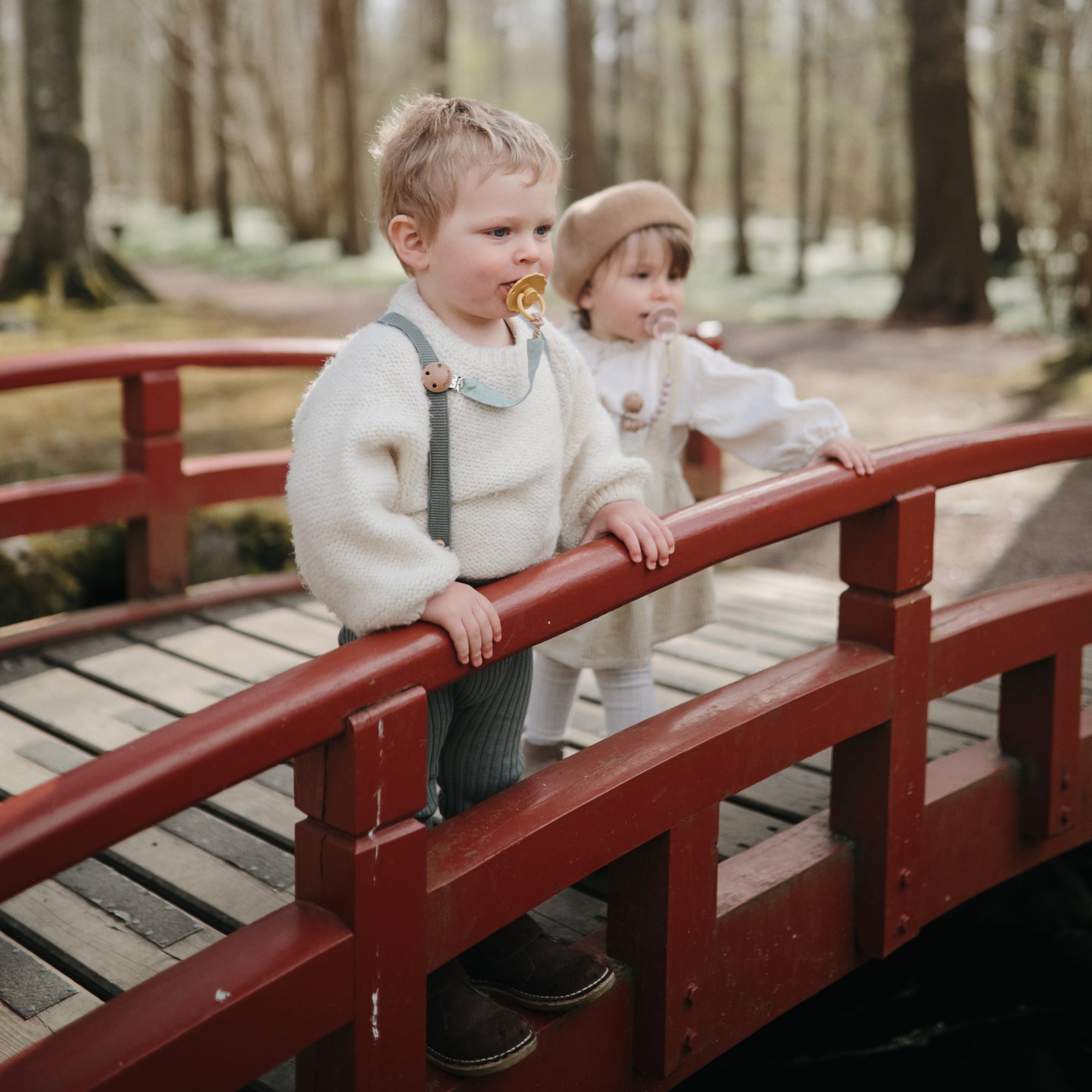 Two kids with FRIGG Daisy Natural Rubber Pacifiers stand on a red bridge in the forest.