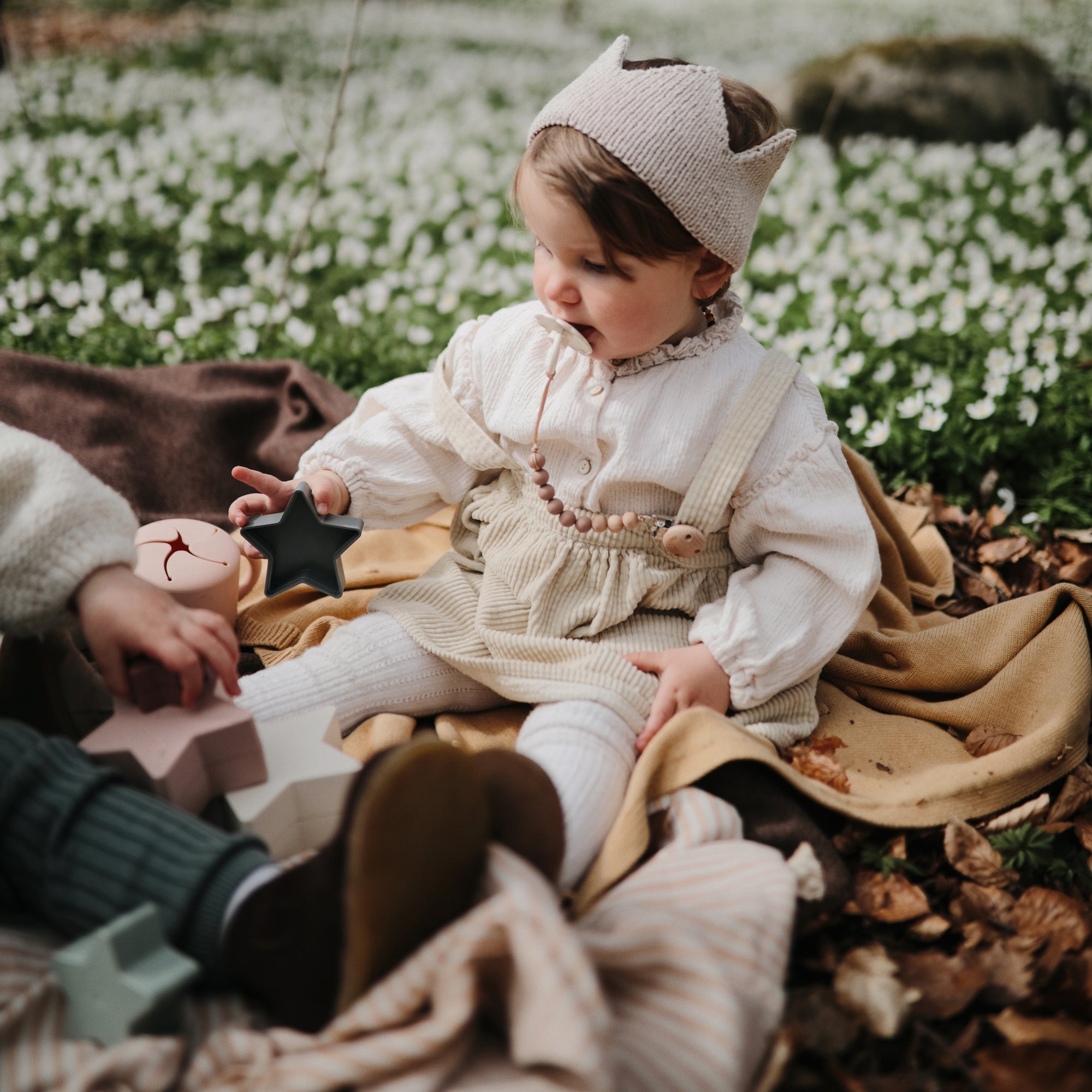 Baby with a crown headband uses FRIGG Daisy Natural Rubber Pacifier while playing on a blanket.