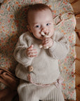 Baby in beige knit outfit with mushie Figurine Teether, lying in floral bassinet.