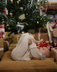 Baby with FRIGG Rope pacifier by Mushie lies on cushion near gifts under a Christmas tree.