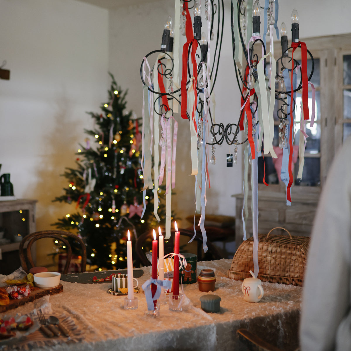 A table with mushie Stacking Cups, candles, and ribbons, a Christmas tree and lights behind.