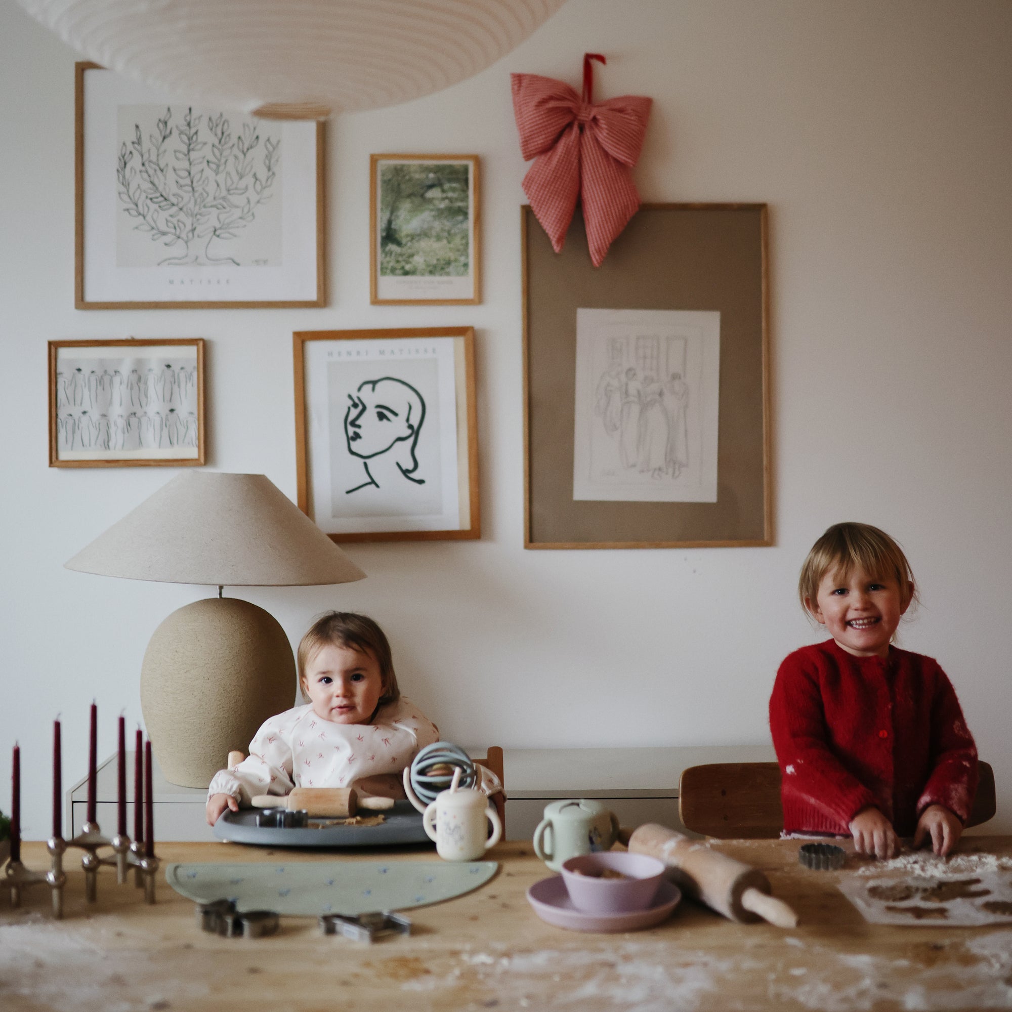 Two smiling children sit at a table with baking supplies, dough, and a mushie Bluey x Mushie Snack Cup. Behind them, framed art and a large red bow decorate the wall as they enjoy baking together.