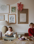 Two smiling children sit at a table with baking supplies, dough, and a mushie Bluey x Mushie Snack Cup. Behind them, framed art and a large red bow decorate the wall as they enjoy baking together.