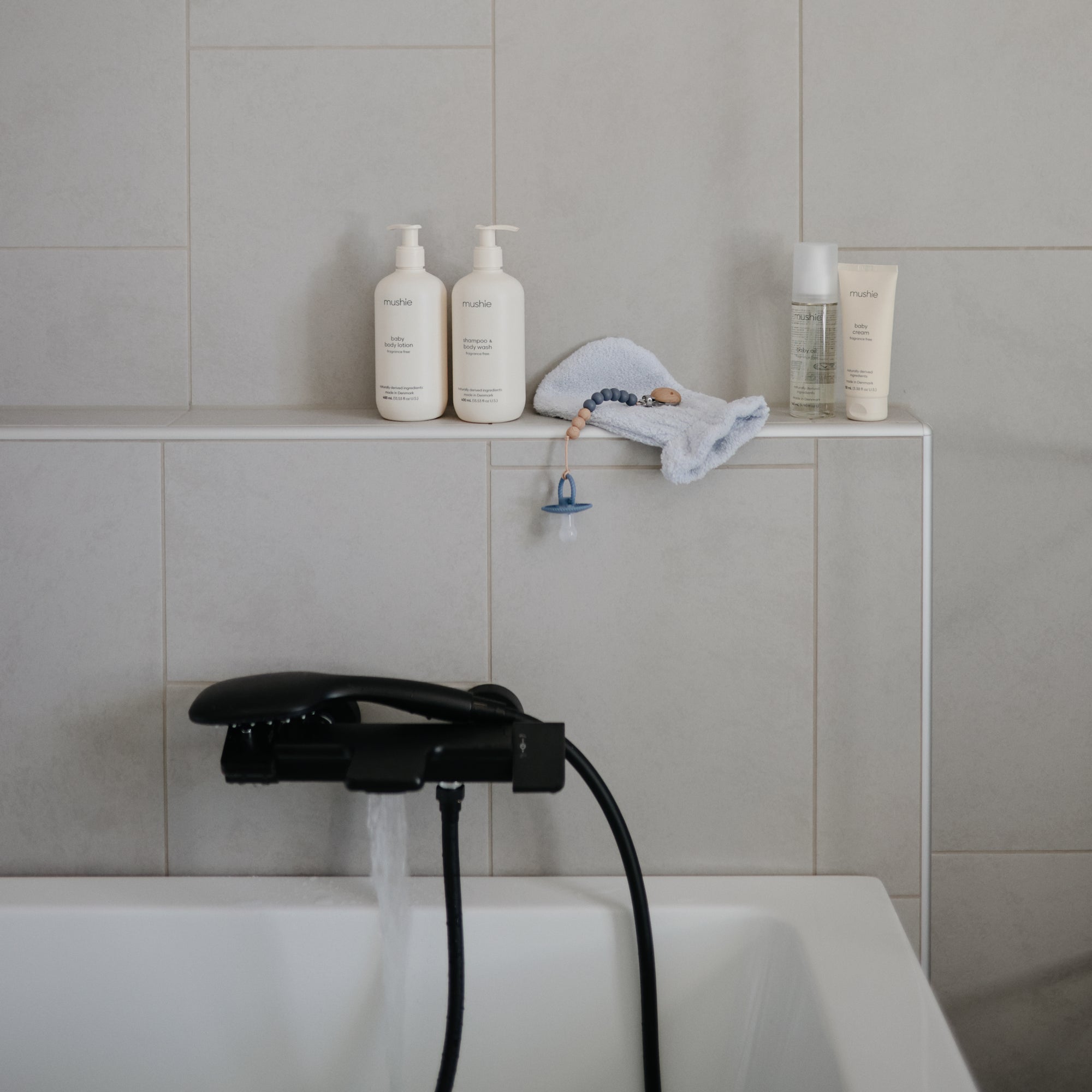 A modern bathtub with water running from a black faucet. On the ledge are skincare products, a white bath mitt, a small spray bottle, and Mushie’s FRIGG Rope Natural Rubber Pacifier 2-Pack.