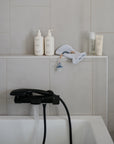 A modern bathtub with water running from a black faucet. On the ledge are skincare products, a white bath mitt, a small spray bottle, and Mushie’s FRIGG Rope Natural Rubber Pacifier 2-Pack.