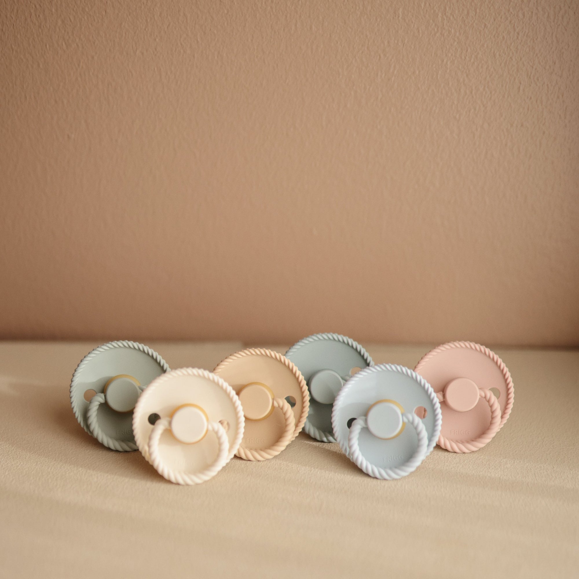 Six FRIGG Rope Natural Rubber Pacifier 2-Packs by Mushie, featuring pastel peach, beige, and light blue shades, are arranged on a soft cream surface against a beige wall.