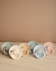 Six FRIGG Rope Natural Rubber Pacifier 2-Packs by Mushie, featuring pastel peach, beige, and light blue shades, are arranged on a soft cream surface against a beige wall.