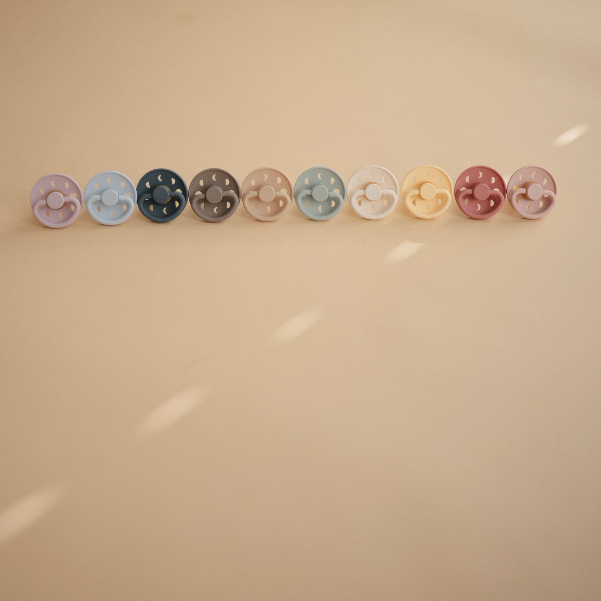 Five FRIGG Moon Natural Rubber Pacifier 2-Packs from FRIGG, featuring assorted colors, are arranged in a line on a beige surface under soft lighting with gentle shadows—an essential for any baby.