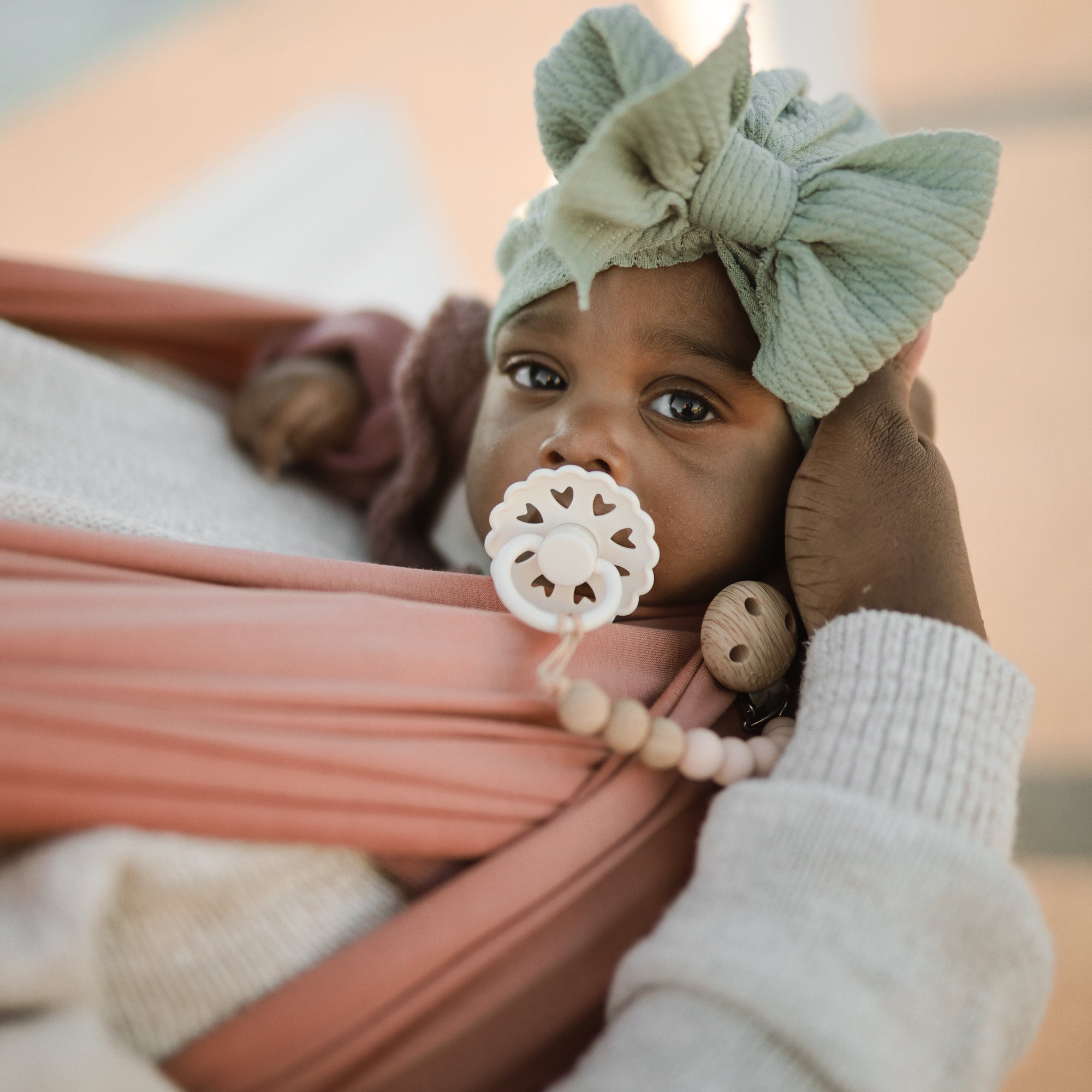 A baby with a green head wrap and a FRIGG Andersen Fairytale Natural Rubber Pacifier is gently held in an adult’s hand, wrapped in a pink sling and gazing upward with wide eyes.