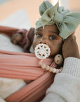 A baby with a green head wrap and a FRIGG Andersen Fairytale Natural Rubber Pacifier is gently held in an adult’s hand, wrapped in a pink sling and gazing upward with wide eyes.