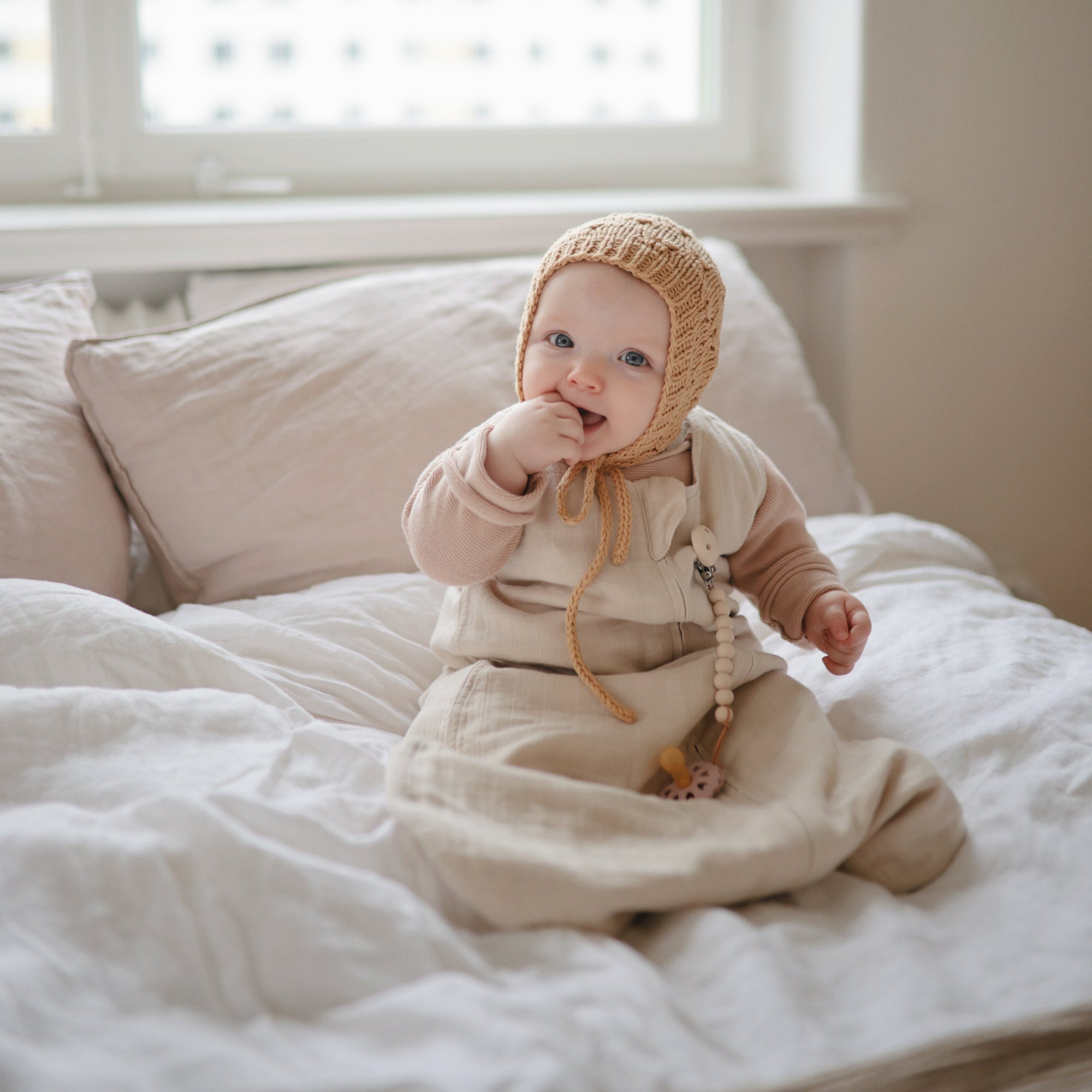 A baby in a beige outfit and knitted hat sits on white bedding, smiling and holding a FRIGG Andersen Fairytale Natural Rubber Pacifier 2-Pack by FRIGG, with soft natural light streaming in from the window behind.