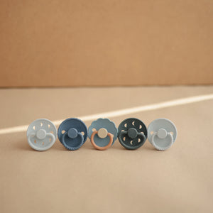 Lifestyle image of a blue-themed line of Frigg pacifiers, featuring the Powder Blue and Slate Moon Pacifiers. 