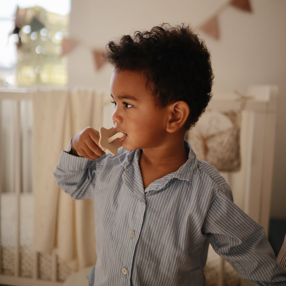Lifestyle image of a young toddler boy with dark curls and blue striped pajamas holding a Shifting Sand Star Training Toothbrush and brushing his teeth