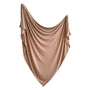 A large, beige Stretchy Swaddle by mushie, made from silky breathable fabric, is draped in elegant, swagged folds against a white background.