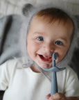 Lifestyle image of a young toddler boy wearing fuzzy bear ear bonnet laying down and smiling into the camera holding a Tradewinds Star Training Toothbrush