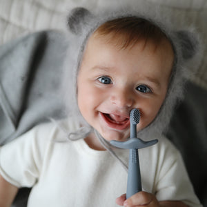 Lifestyle image of a young toddler boy wearing fuzzy bear ear bonnet laying down and smiling into the camera holding a Tradewinds Star Training Toothbrush